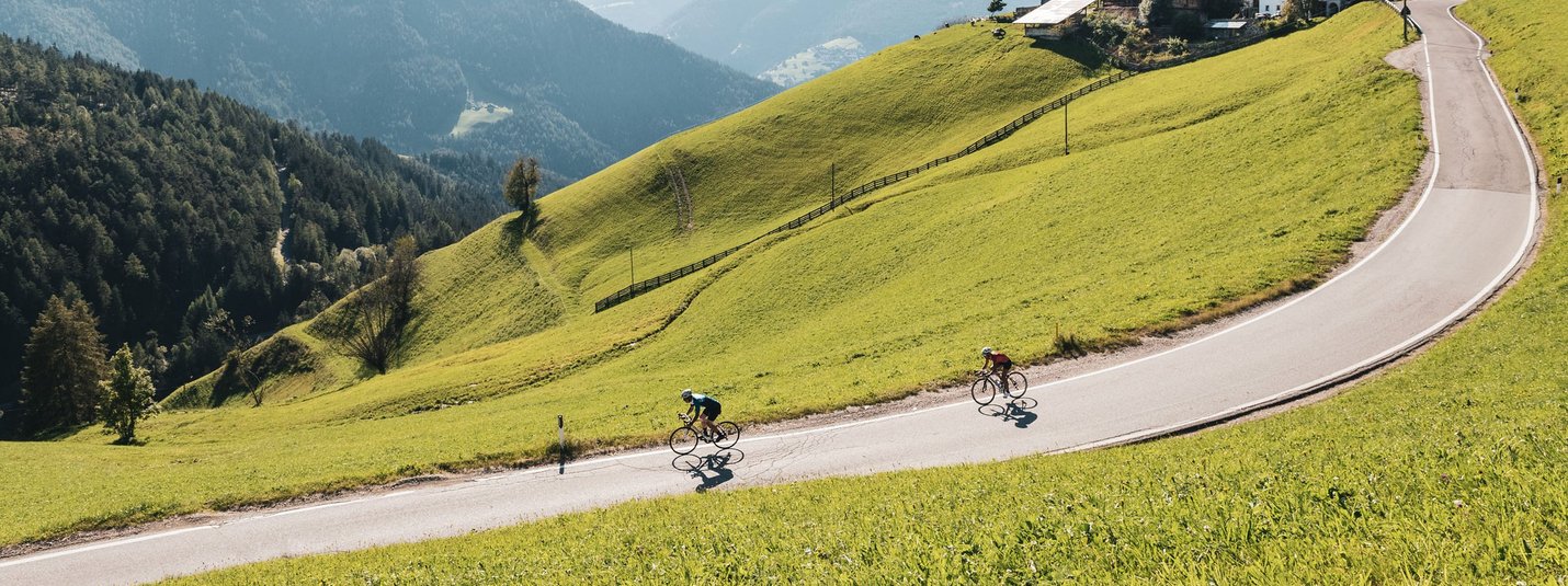 Zwei Radfahrer fahren eine kurvige Bergstraße umgeben von grünen Hügeln