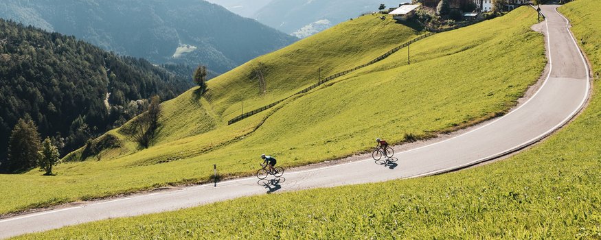 Two cyclists ride a winding mountain road surrounded by green hills