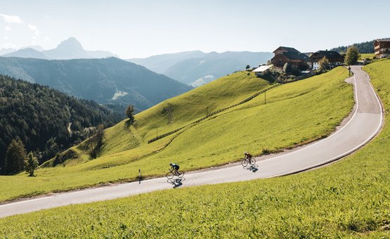 Two cyclists ride a winding mountain road surrounded by green hills