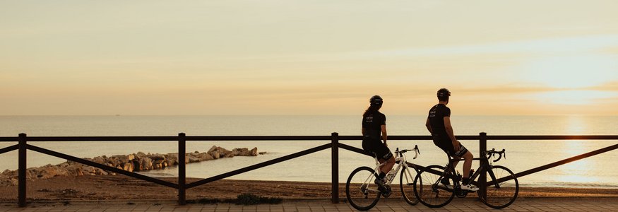 Zwei Radfahrer blicken bei Sonnenuntergang aufs Meer am Promenadenweg