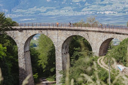Cyclists riding on a stone arched bridge over a forested valley