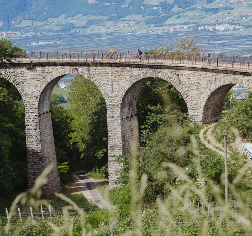 Kurven, Kultur & Kontraste in Südtirols Süden © Thomas Monsorno - TV Castelfeder Fahrradfahrer auf einer Steinbrücke mit Bögen in einer grünen Landschaft