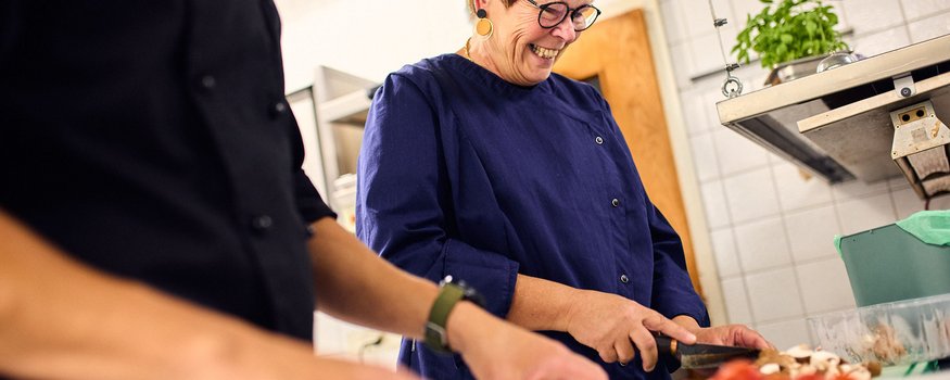 Frau schneidet Gemüse in professioneller Küche, lächelnd und konzentriert