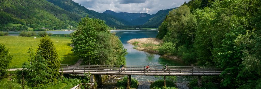 Three cyclists crossing a bridge over a river in a mountainous forest landscape