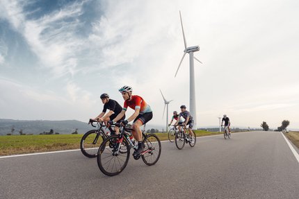 Cinque ciclisti pedalano su una strada vicino a turbine eoliche