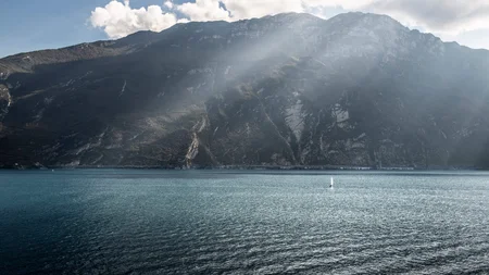 Zwischen Passstraßen, See & Dolce Vita beim Rennradurlaub am nördlichen Gardasee © Alex Moling Berg mit Sonnenstrahlen über blauem See und kleinem Segelboot