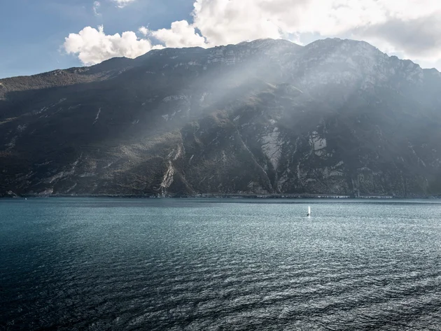 Zwischen Passstraßen, See & Dolce Vita beim Rennradurlaub am nördlichen Gardasee © Alex Moling Berg mit Sonnenstrahlen über blauem See und kleinem Segelboot