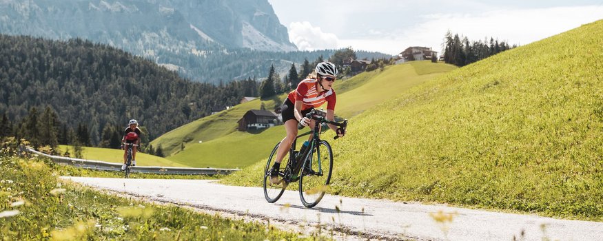 San Vigilio-Pässe-Quartett © Alex Moling Zwei Radfahrer fahren auf Bergstraße mit Gipfel in der Sonne