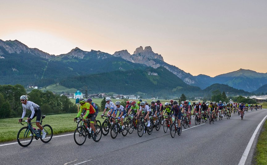 Large group of cyclists riding on a rural road with mountains behind