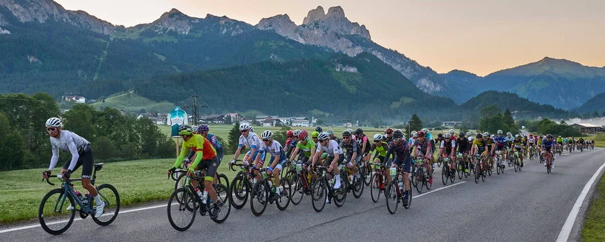 Large group of cyclists riding on a rural road with mountains behind