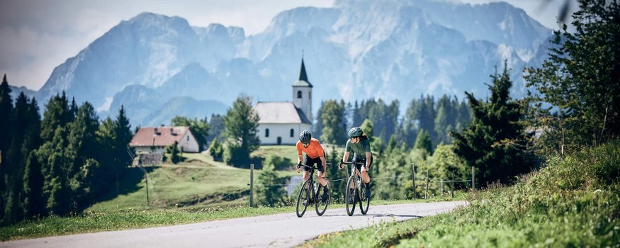 Zwei Fahrradfahrer auf Landstraße vor Kirche und Bergen in grüner Landschaft