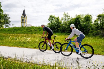 Mittelkärnten © Michael Stabentheiner Zwei Radfahrer fahren auf Landstraße, Kirche und grüne Wiese im Hintergrund