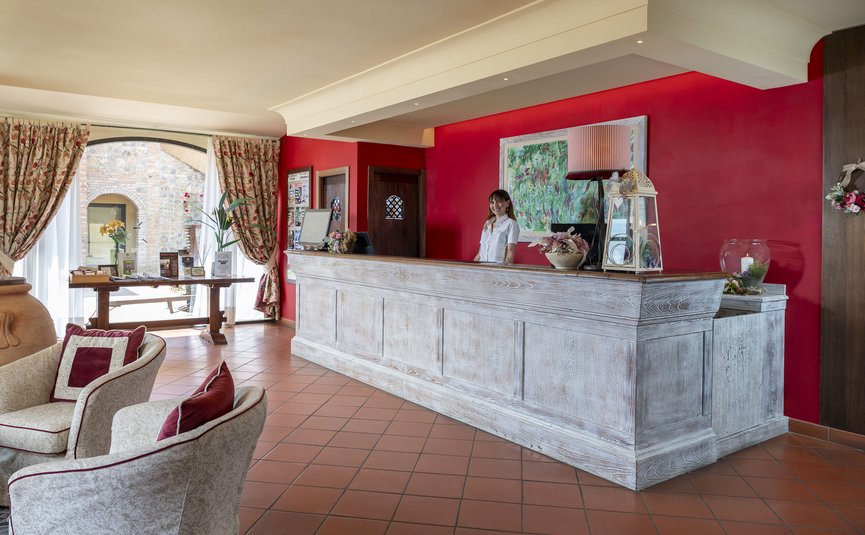Hotel & Restaurant Casolare Le Terre Rosse ***S © Hotel Casolare Le Terre Rosse Hotelrezeption mit einer Mitarbeiterin an einem weißen hölzernen Empfangstresen
