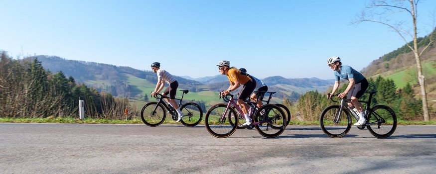 Vier Radfahrer fahren auf Landstraße mit Bergen und blauem Himmel im Hintergrund