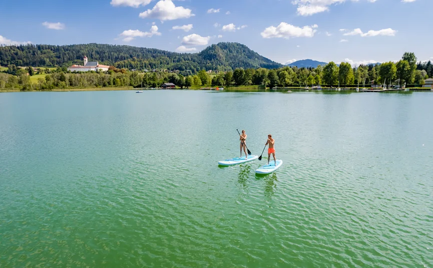 Zwei Personen paddleboarden auf einem See vor bewaldeten Hügeln und blauem Himmel