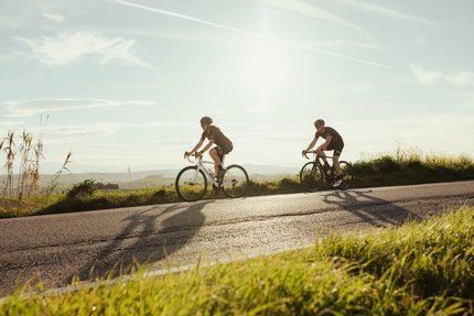 Zwei Radfahrer fahren auf einer Straße in ländlicher Umgebung bei Sonnenschein
