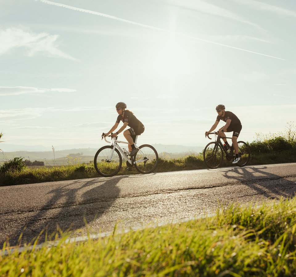 Zwei Radfahrer fahren auf einer Straße in ländlicher Umgebung bei Sonnenschein