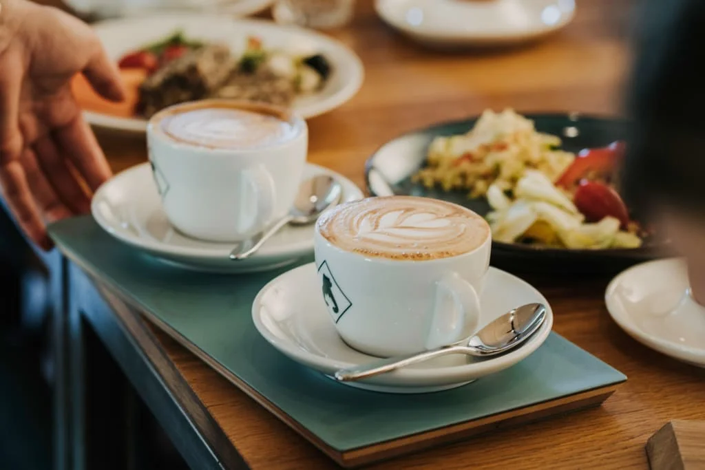 Last Minute 15% Rabatt zwei tasse cappuccino mit milchglasur auf einem holztisch neben einem gericht mit lebensmitteln