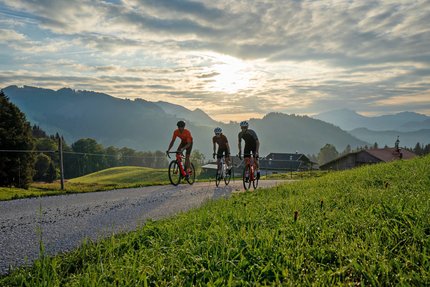 Tre ciclisti pedalano su strada di campagna con montagne al tramonto.
