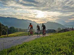 Tre ciclisti pedalano su strada di campagna con montagne al tramonto.