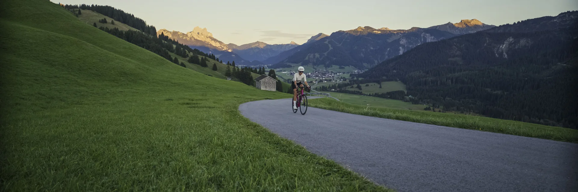 Radfahrer auf Bergstraße mit Grünfläche und Bergkulisse bei Sonnenuntergang