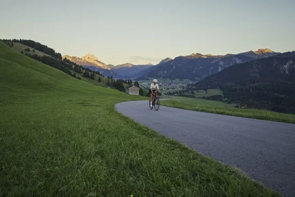 Ciclista su strada di montagna con campi verdi e vista sulle montagne al tramonto