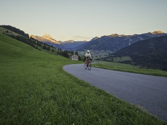 Radfahrer auf Bergstraße mit Grünfläche und Bergkulisse bei Sonnenuntergang