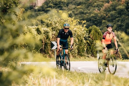Due ciclisti su strada davanti a un castello in paesaggio boschivo