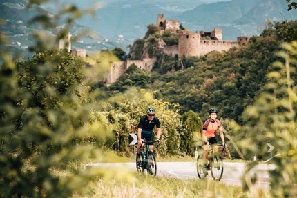 Zwei Radfahrer auf Straße vor einer Burg in bewaldeter Landschaft