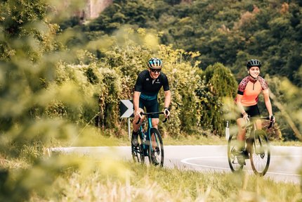 Zwei Radfahrer auf Straße vor einer Burg in bewaldeter Landschaft