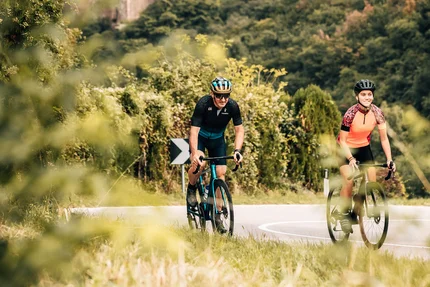 Due ciclisti su strada davanti a un castello in paesaggio boschivo
