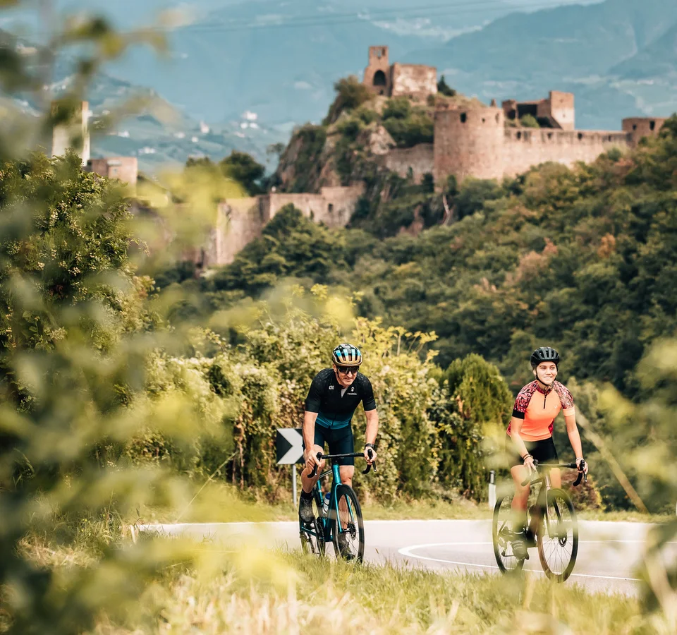 Golden autumn on the road bike in Tramin © Jens Scheibe Two cyclists on road with castle in forested landscape background