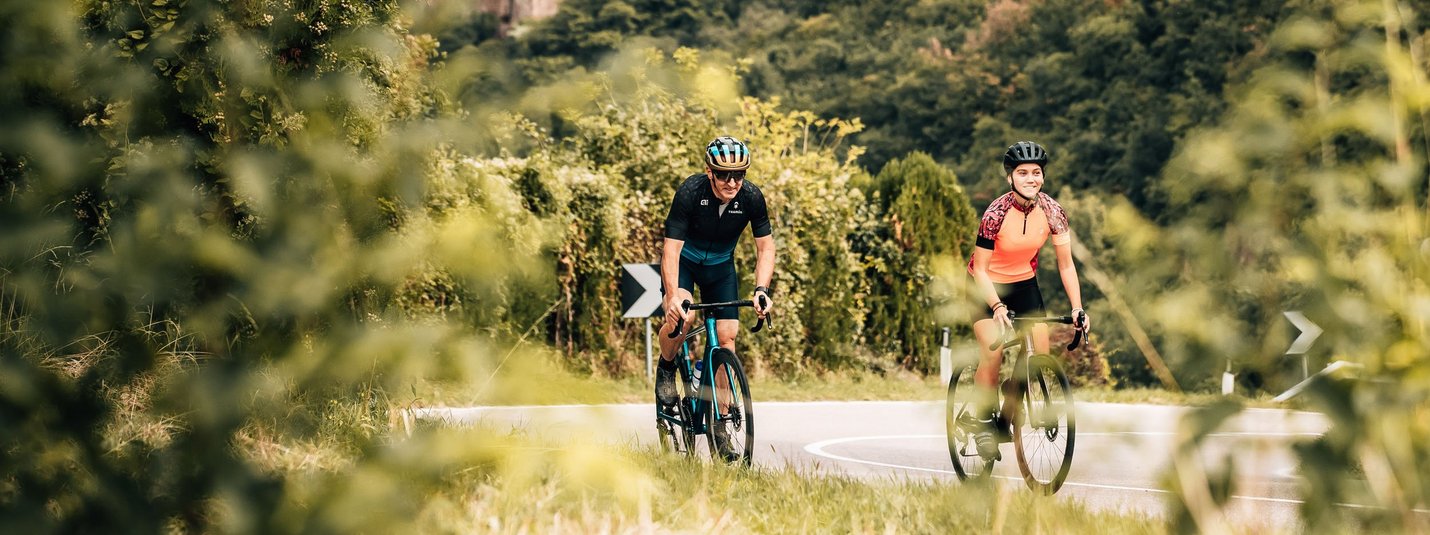 Etschtalrunde © Jens Scheibe Zwei Radfahrer auf Straße vor einer Burg in bewaldeter Landschaft