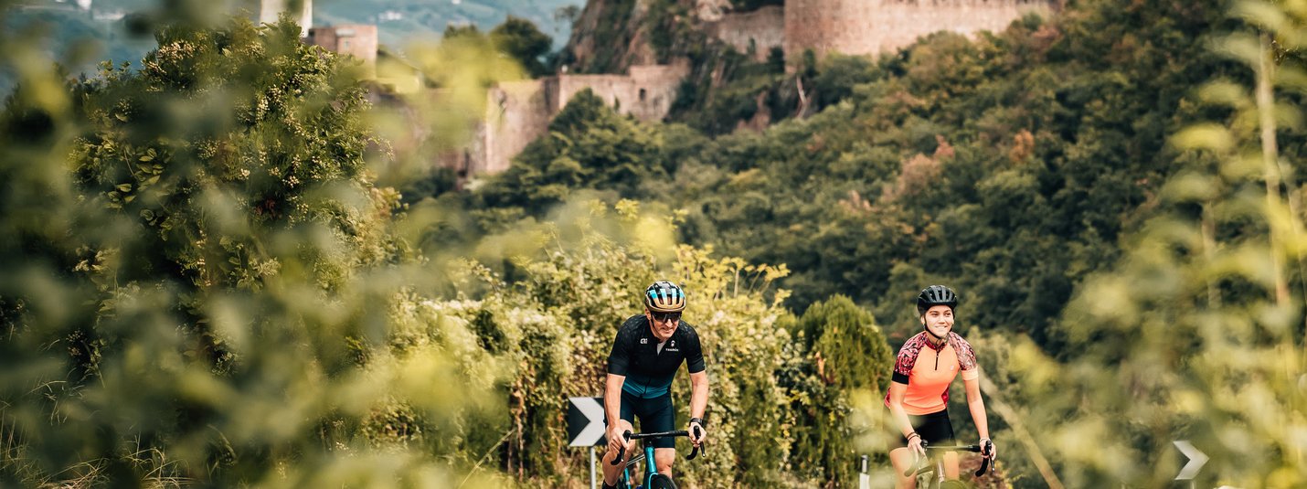 Etschtalrunde © Jens Scheibe Zwei Radfahrer auf Straße vor einer Burg in bewaldeter Landschaft