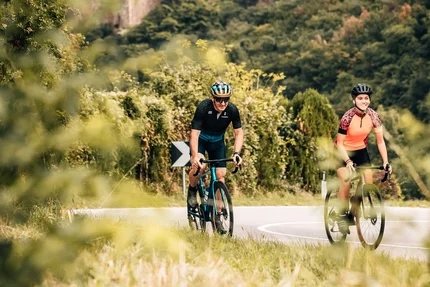 Zwei Radfahrer auf Straße vor einer Burg in bewaldeter Landschaft