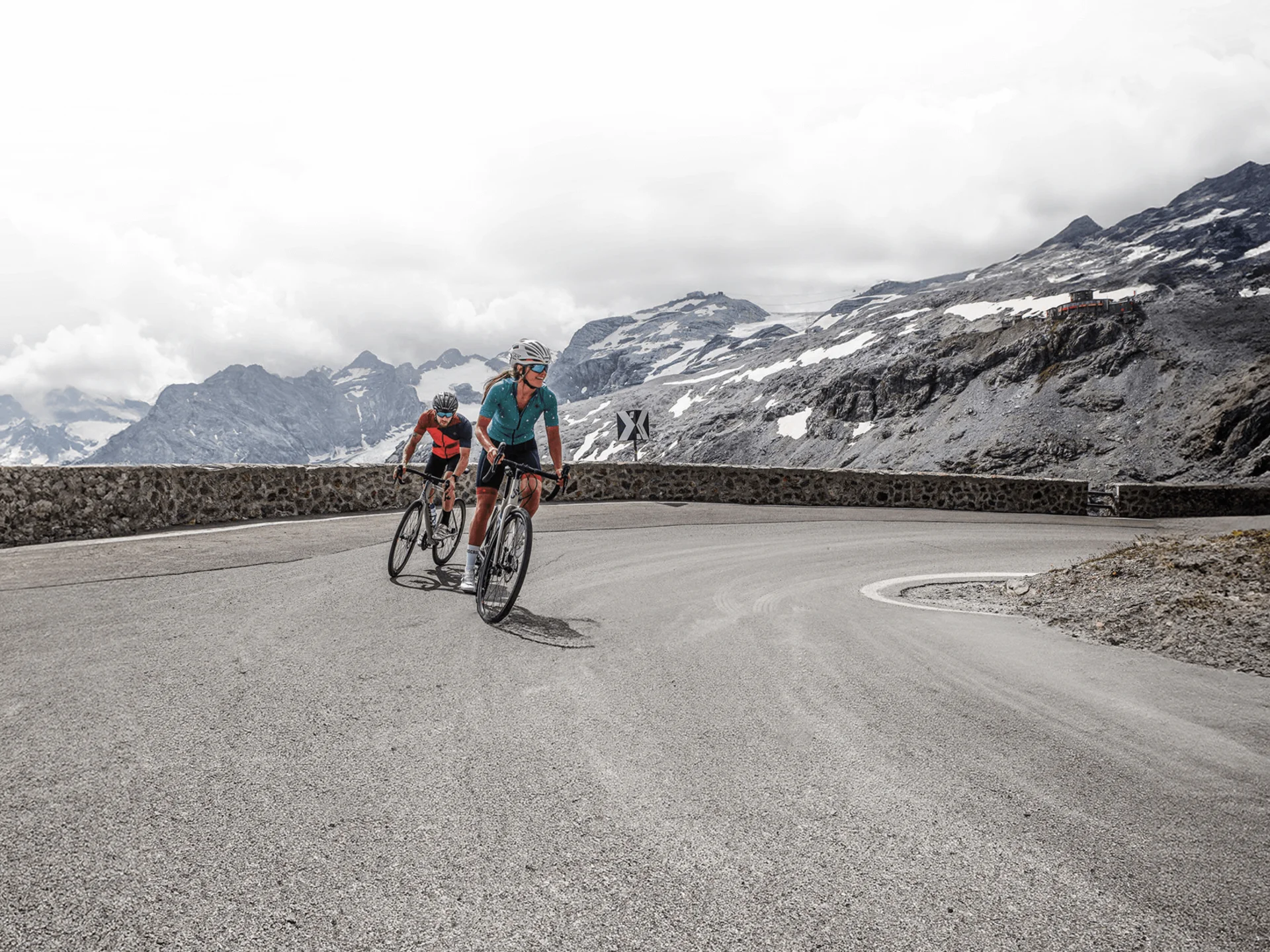 Due ciclisti su una strada montana tortuosa con cime innevate sullo sfondo