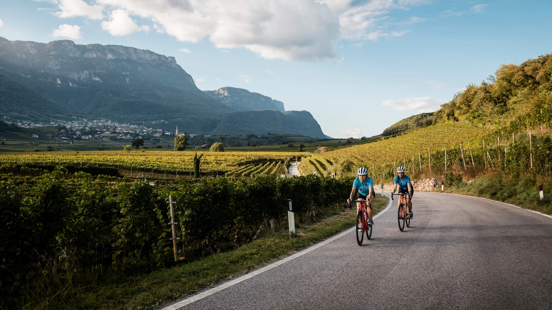 Golden autumn on the road bike in Tramin Two cyclists on rural road through vineyards with mountains in background