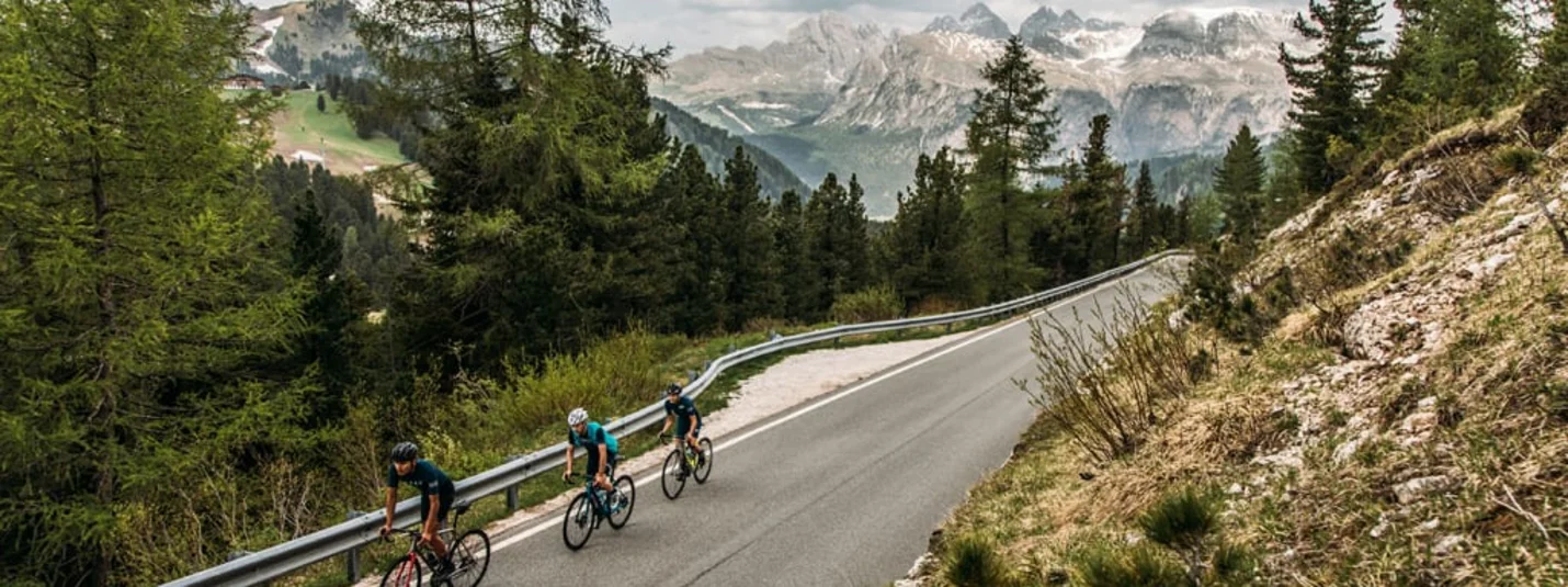 Dolomites Road Bike Week Tre ciclisti su una strada di montagna con pini e montagne sullo sfondo