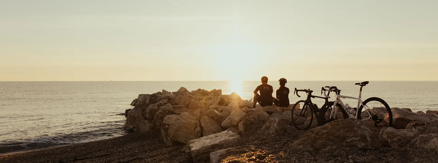 Gevoelens van opgetogenheid bij de zee en het meer © Alex Moling Twee fietsers zitten op rotsen aan het strand en kijken naar de zonsondergang