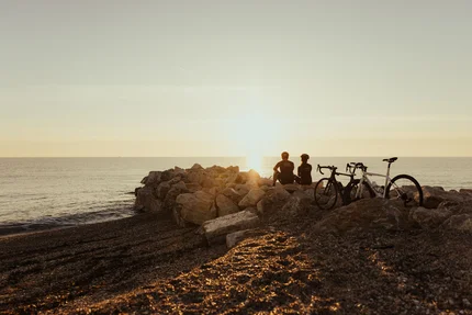 Zwei Radfahrer sitzen auf Felsen am Strand und schauen auf den Sonnenuntergang