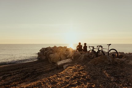 Due ciclisti seduti sulle rocce in spiaggia a guardare il tramonto