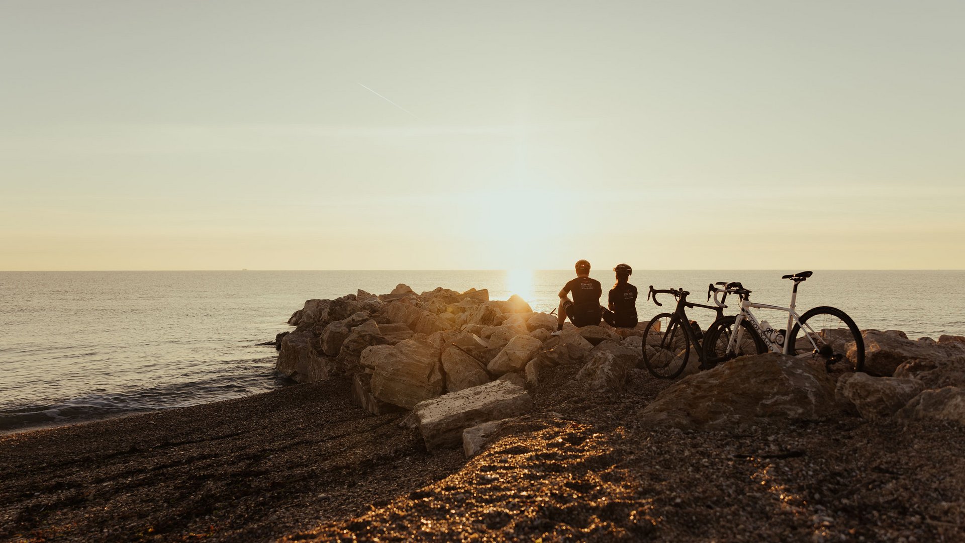 Zwei Radfahrer sitzen auf Felsen am Strand und schauen auf den Sonnenuntergang