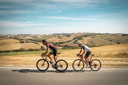 Due ciclisti pedalano su strada di campagna tra colline ondulate.