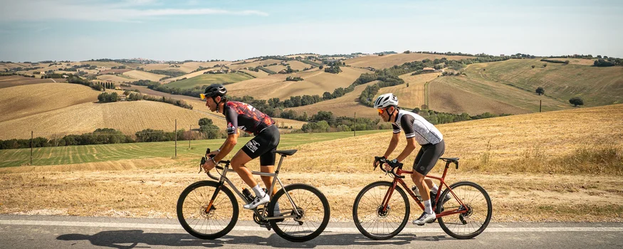 Zwei Radfahrer fahren auf Landstraße durch hügelige Landschaft.