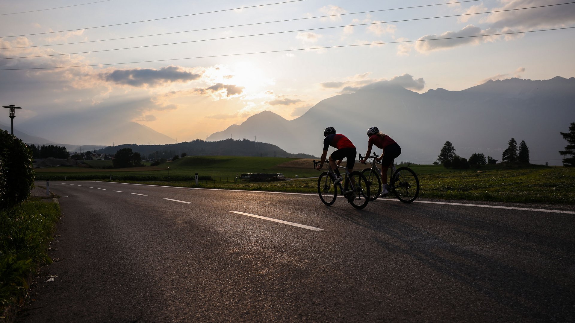 Two cyclists riding on a road at sunset with mountains in the background