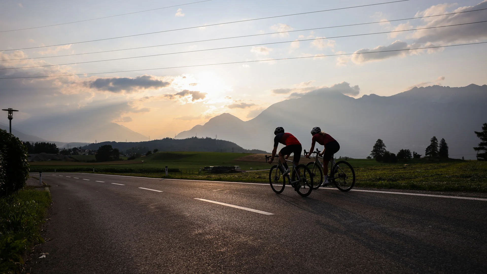 Due ciclisti pedalano su una strada al tramonto con montagne sullo sfondo