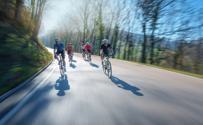 Groep fietsers rijdt snel over een weg door het bos op een zonnige dag