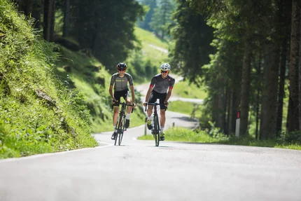 Due ciclisti su una strada di montagna immersa nel bosco in una giornata di sole