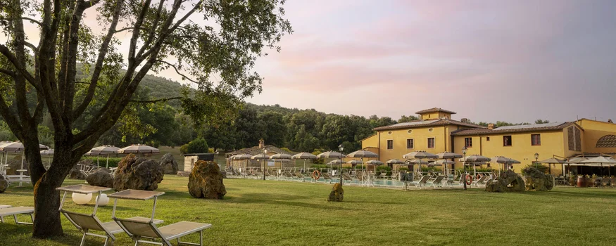 Rennradfahren in der Toskana © Hotel Casolare Le Terre Rosse Grüner Garten mit Bäumen, Liegestühlen und Sonnenschirmen neben einem gelben Gebäude