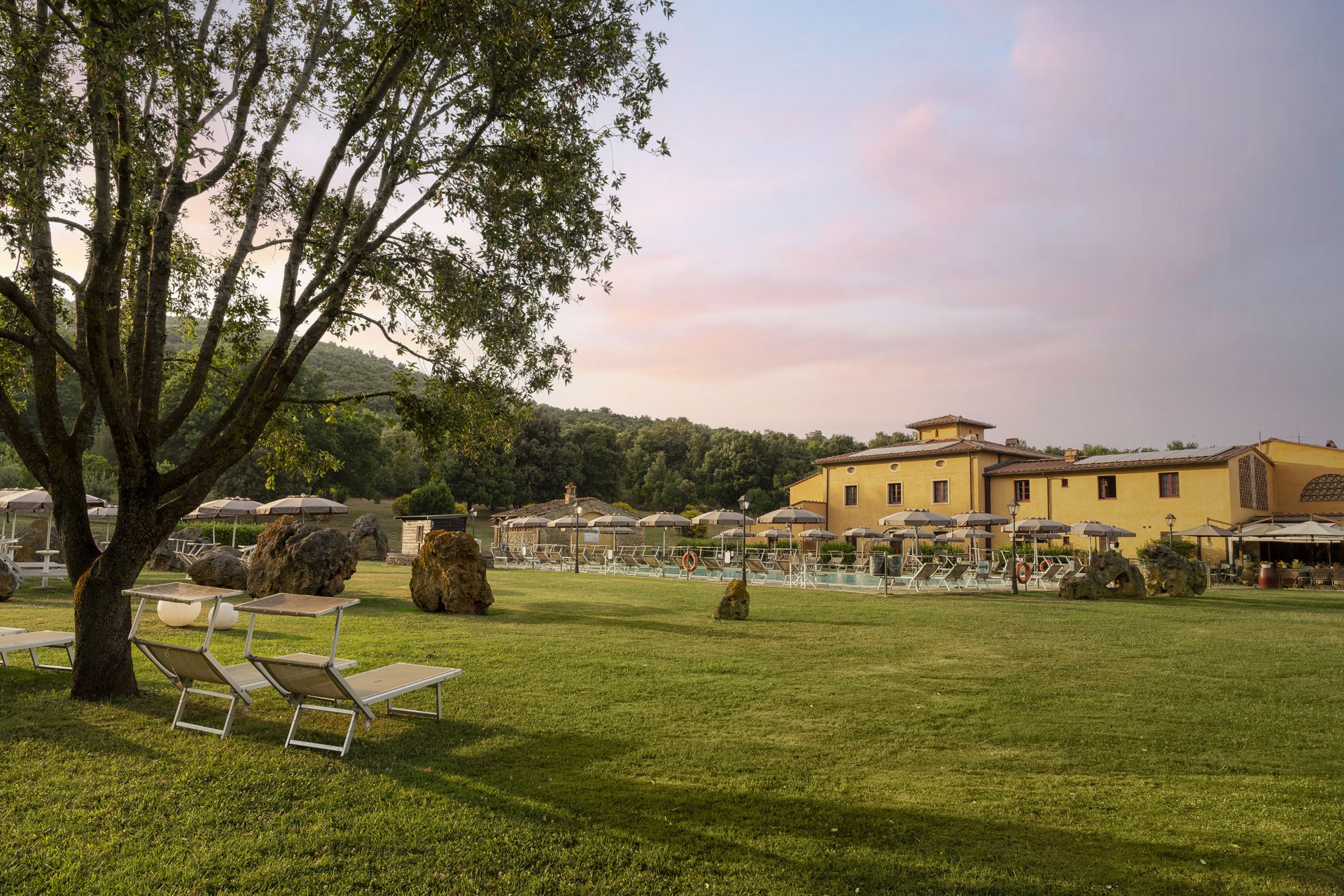 Grüner Garten mit Bäumen, Liegestühlen und Sonnenschirmen neben einem gelben Gebäude